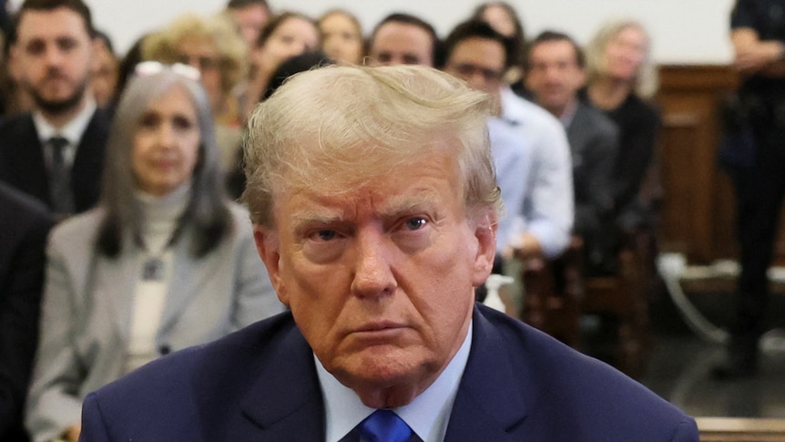 Donald Trump sits in court and looks straight ahead. The gallery behind him is full of people.