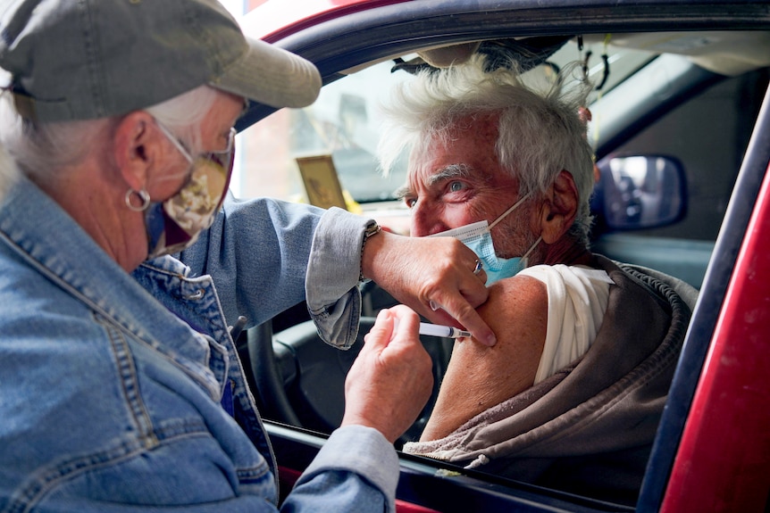 An older man in a mask gets a needle in his shoulder through an open car window