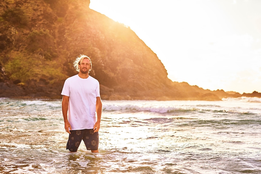 A tall blonde man in a white tshirt and board shorts stands in the waves