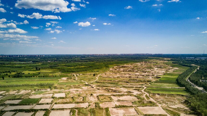 Yonging's dry, sandy riverbed rests among vibrant green fields contrasted with brilliantly blue skies with city in background.