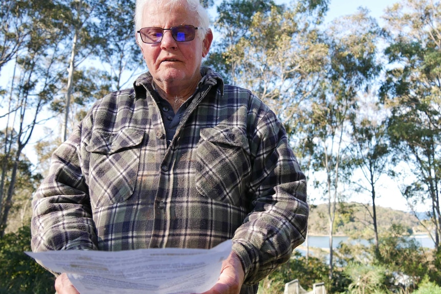 An elderly man wearing transition glasses and a flannel overshirt, pictured in bushland.