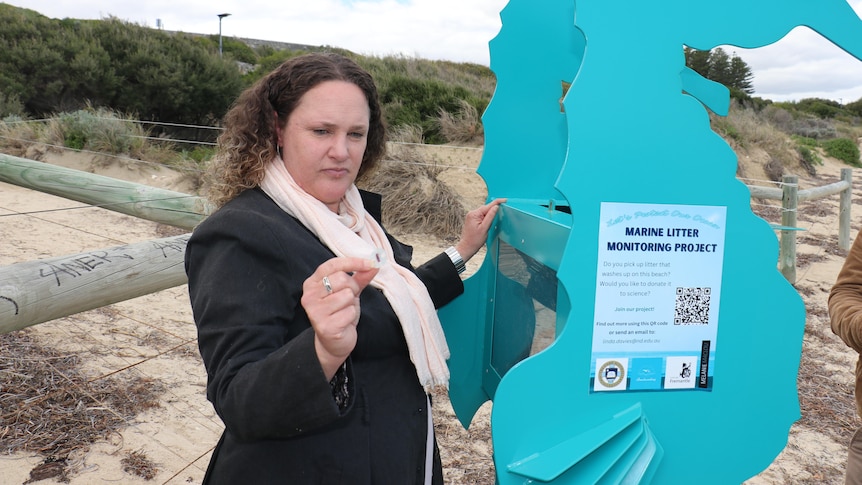 A light blue plastic bin with the design of a seahorse is seen on a beach. Next to it, a woman holds up a piece of rubbish.