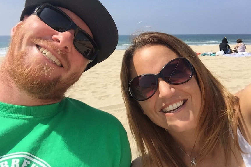 Sandy Casey and a man smile for a selfie on a beach.