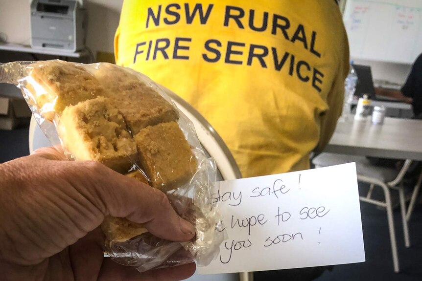 Hand holding home-made cake with note 'Stay Safe', person in RFS uniform in background.