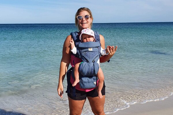A woman with a baby in a baby carrier stands on a beach.