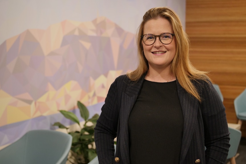 A woman with long blonde hair and glasses smiles at the camera in front of a mountain mural.