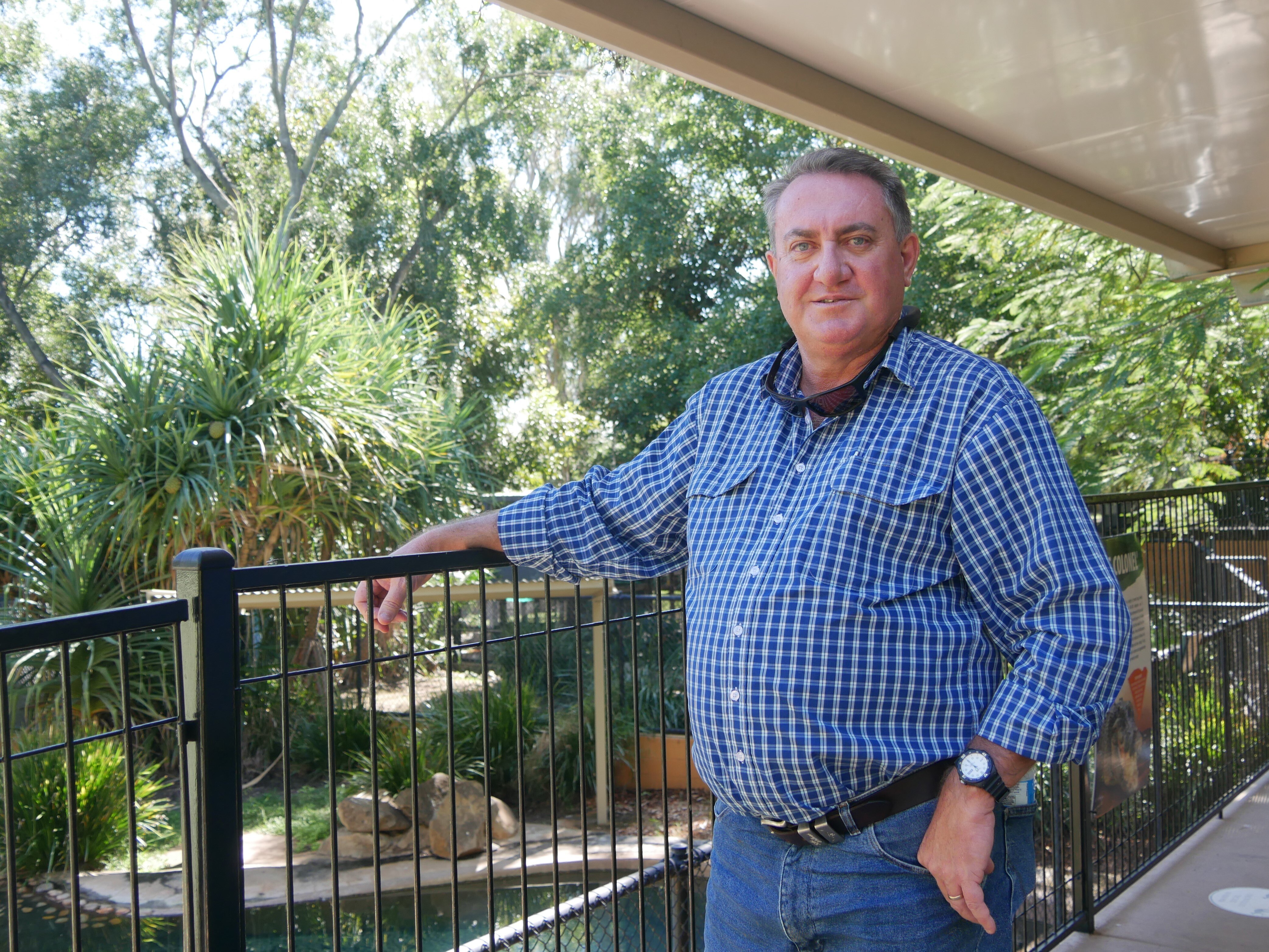 Man in checkered shirt standing against railing with garden and trees in background 