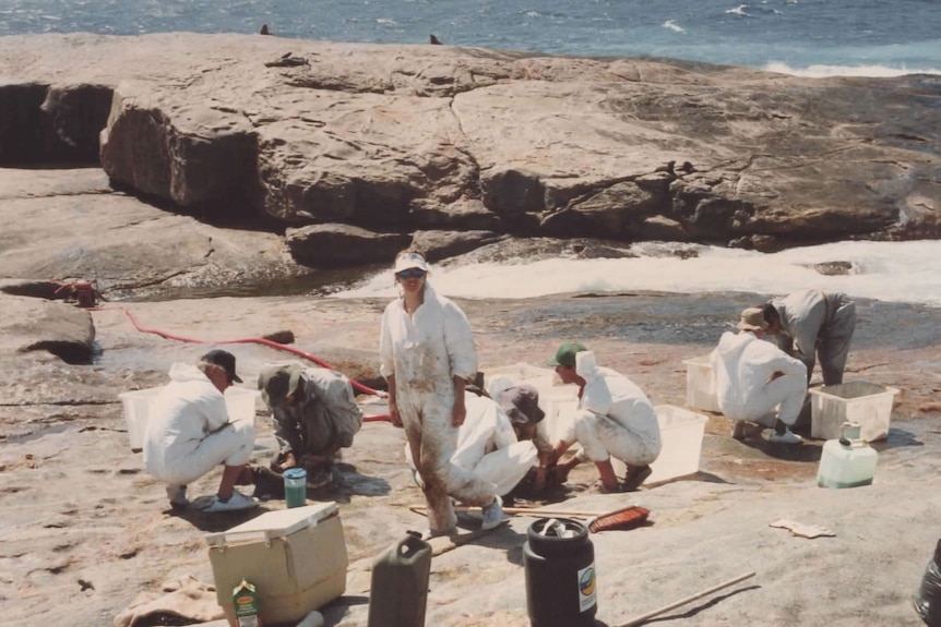 A group of people in white jumpsuits on a rocky plateau fronting the ocean.