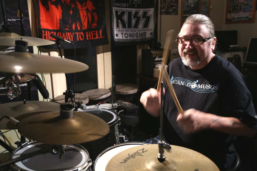 A man sits playing the drums in a room filled with music paraphernalia