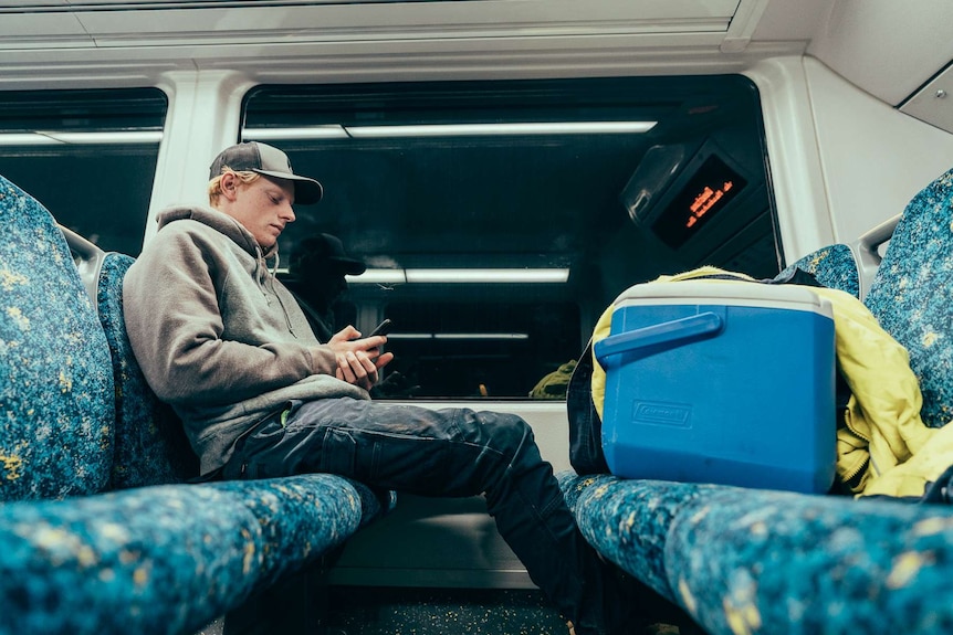 Louis Giles plays on his phone on a train with an esky beside him.