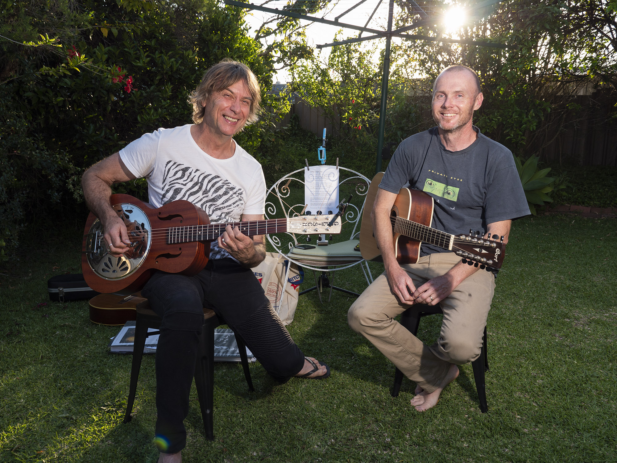 Two males sit in suburban backyard holding guitars as the sun sets.