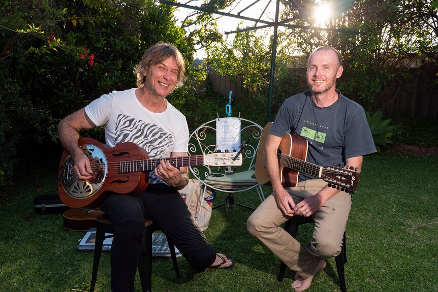 Two males sit in suburban backyard holding guitars as the sun sets