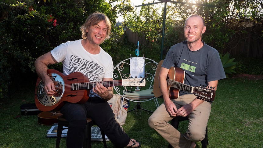 Two males sit in suburban backyard holding guitars as the sun sets.