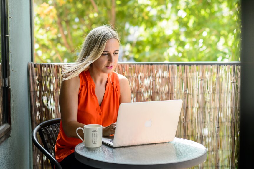 A blonde woman wearing an orange top sits at a table on a balcony, typing on a laptop.