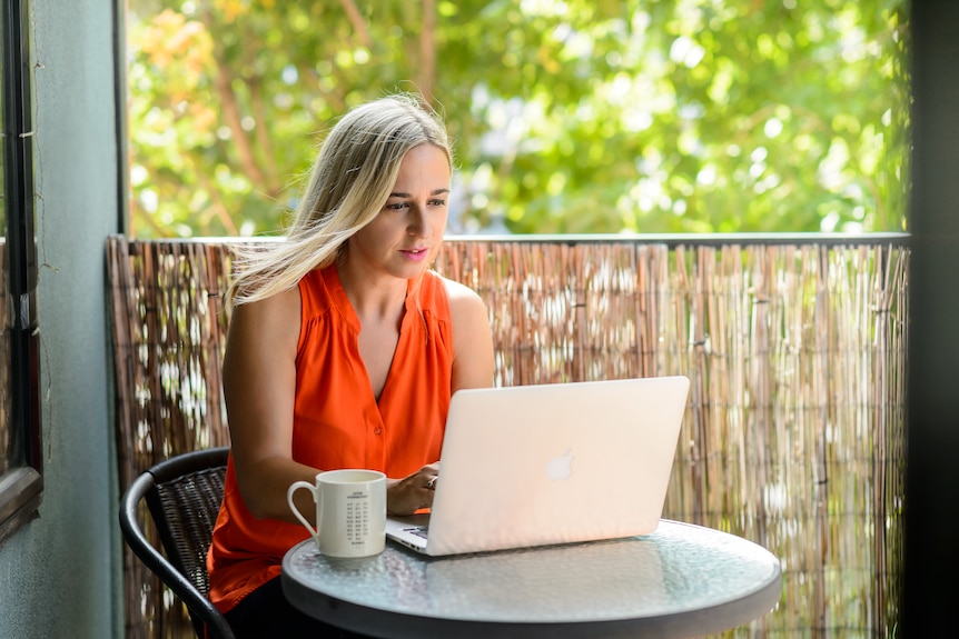 A blonde woman wearing an orange top sits at a table on a balcony, typing on a laptop.