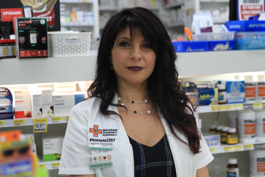 Pharmacist Caroline Diamantis standing at the counter of her chemist with shelves of medicines in the background