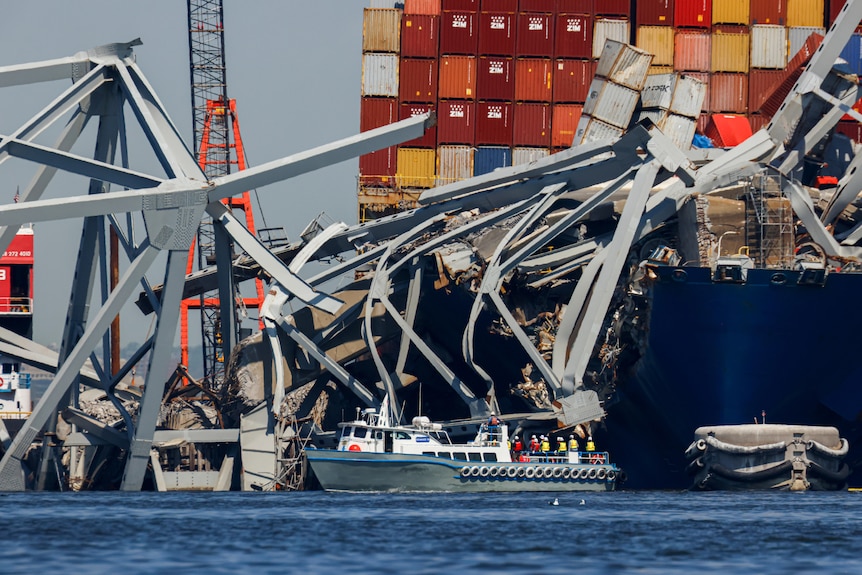 A small boat floats in front of a cargo ship covered in twisted metal.