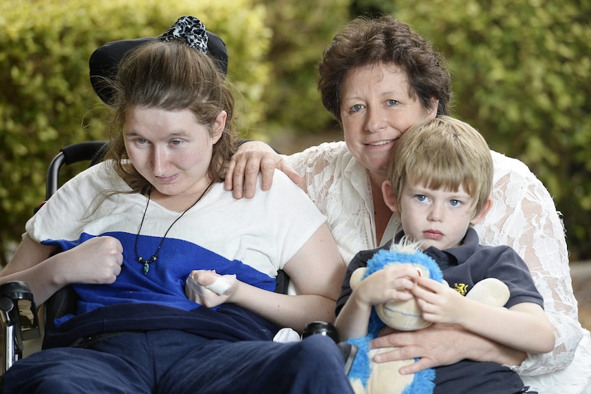 A young woman in a wheelchair looks towards the ground. An older woman with a young boy on her lap sits beside her smiling.