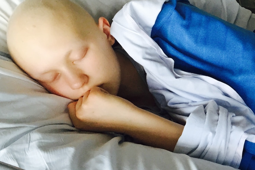 A teenage girl lies asleep on a hospital bed, she is bald from chemotherapy  