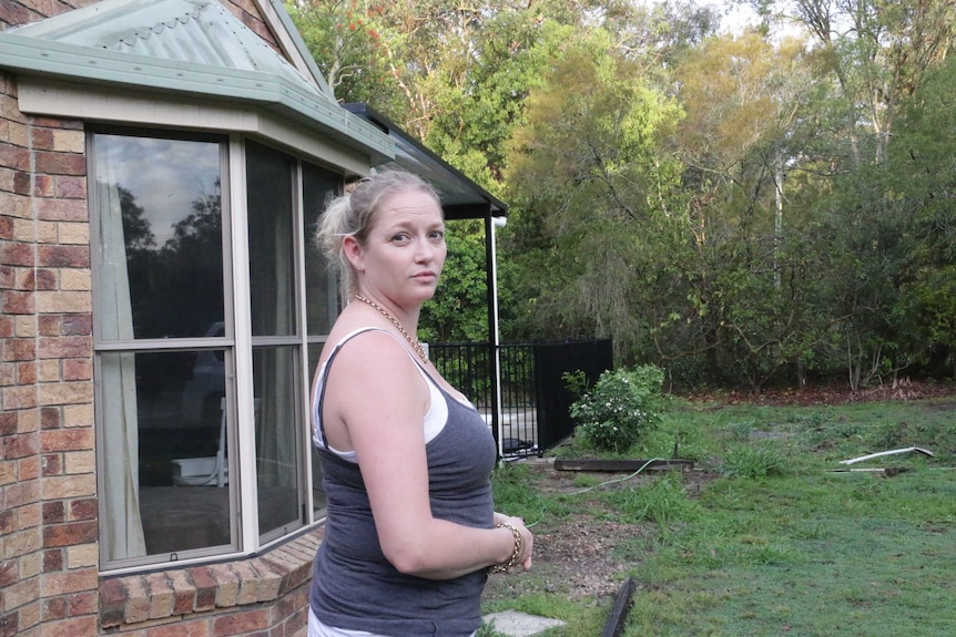 A woman standing on the front step of a one-storey brick house facing the bush