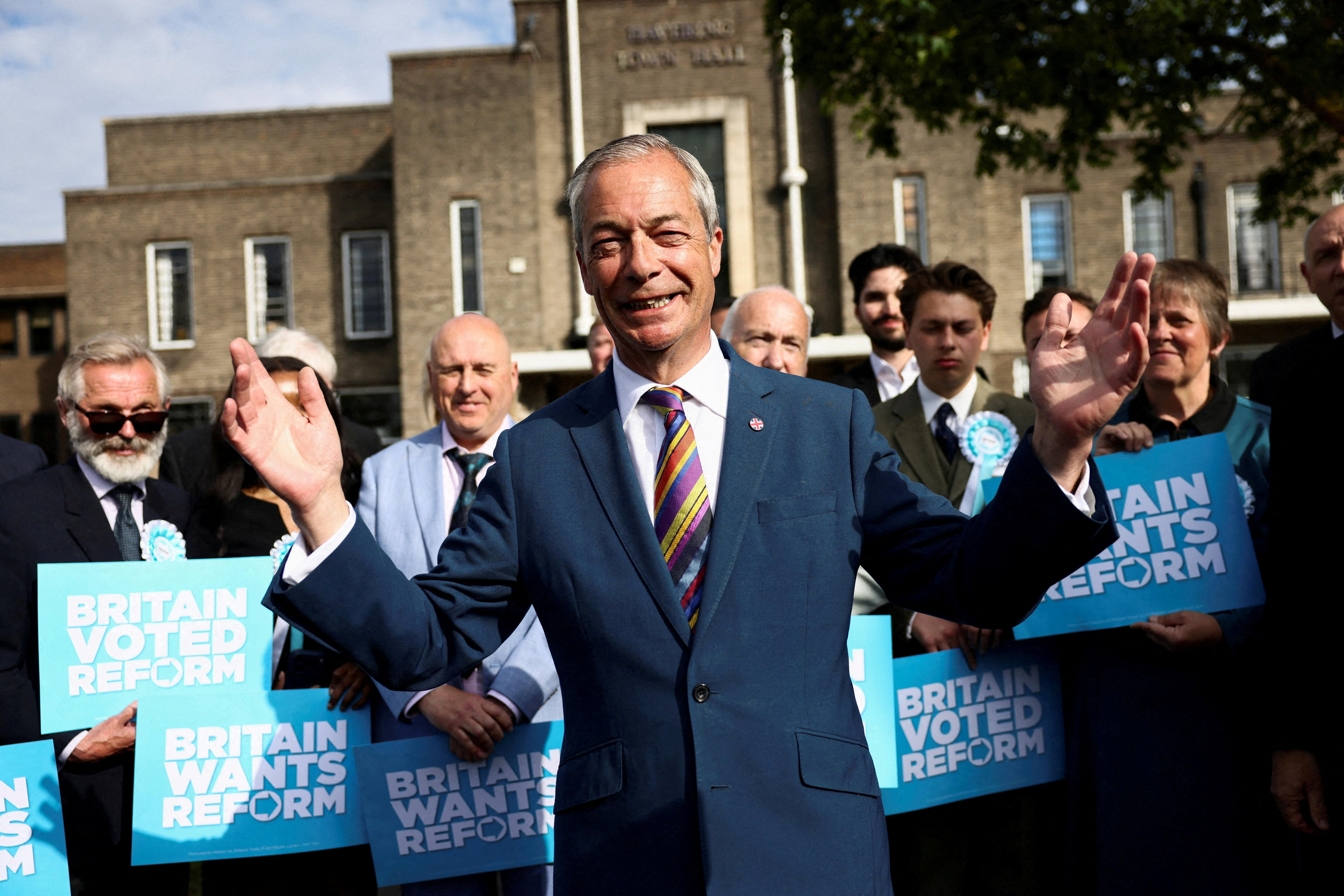 A 60-something man in a blue suit raises his arms in triumph with supporters holding banners behind him on UK street