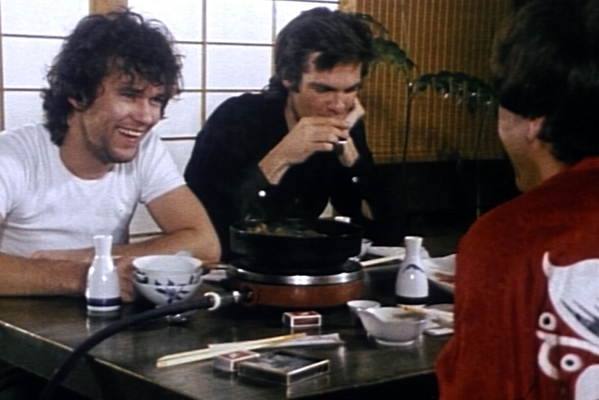 Video still Jimmy Barnes laughing, Don walker sipping tea and back of Molly Meldrum's head. All sit at a low dinner table 
