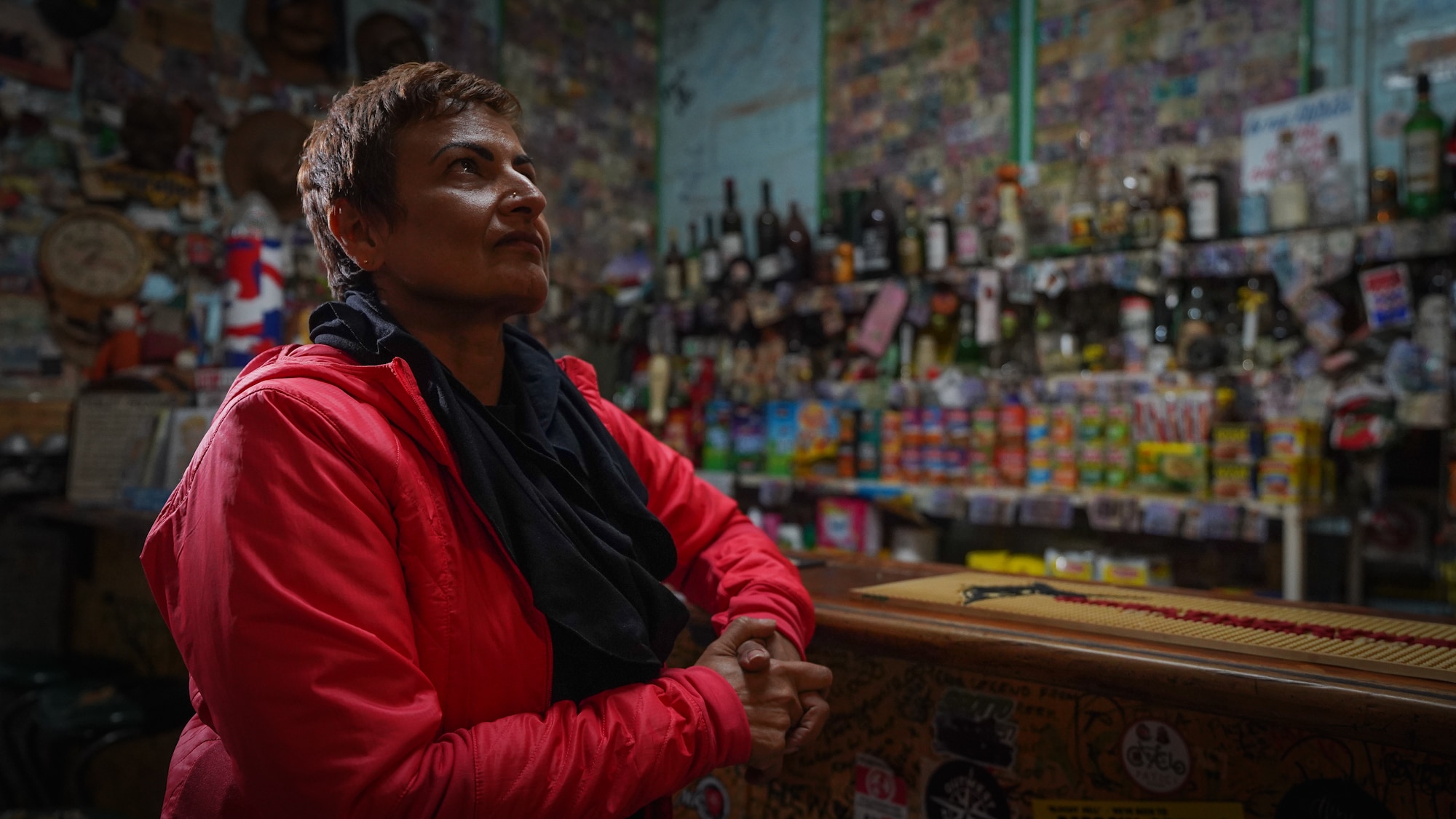 A woman sitting at the bar inside a dim hotel, looking over the mementos plastered on the walls around her.