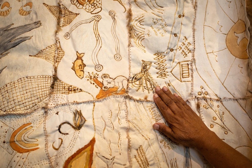 A woman's hand rests on the underside of a possum skin cloak, etched with artwork showing places and animals.
