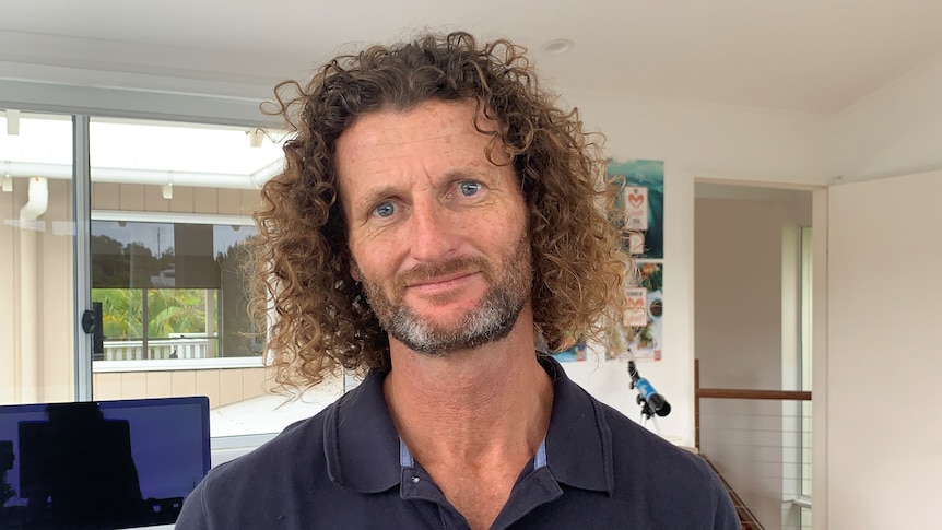A man with shoulder-length curly hair wearing blue shirt looking at the camera in his home office with a computer in background
