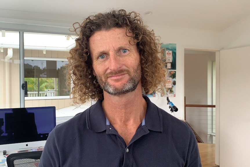 A man with shoulder-length curly hair wearing blue shirt looking at the camera in his home office with a computer in background