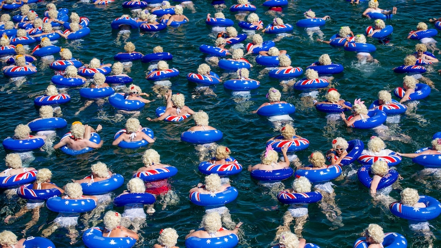 Hundreds of swimmers with blue floaties and blonde wigs swim in the ocean.