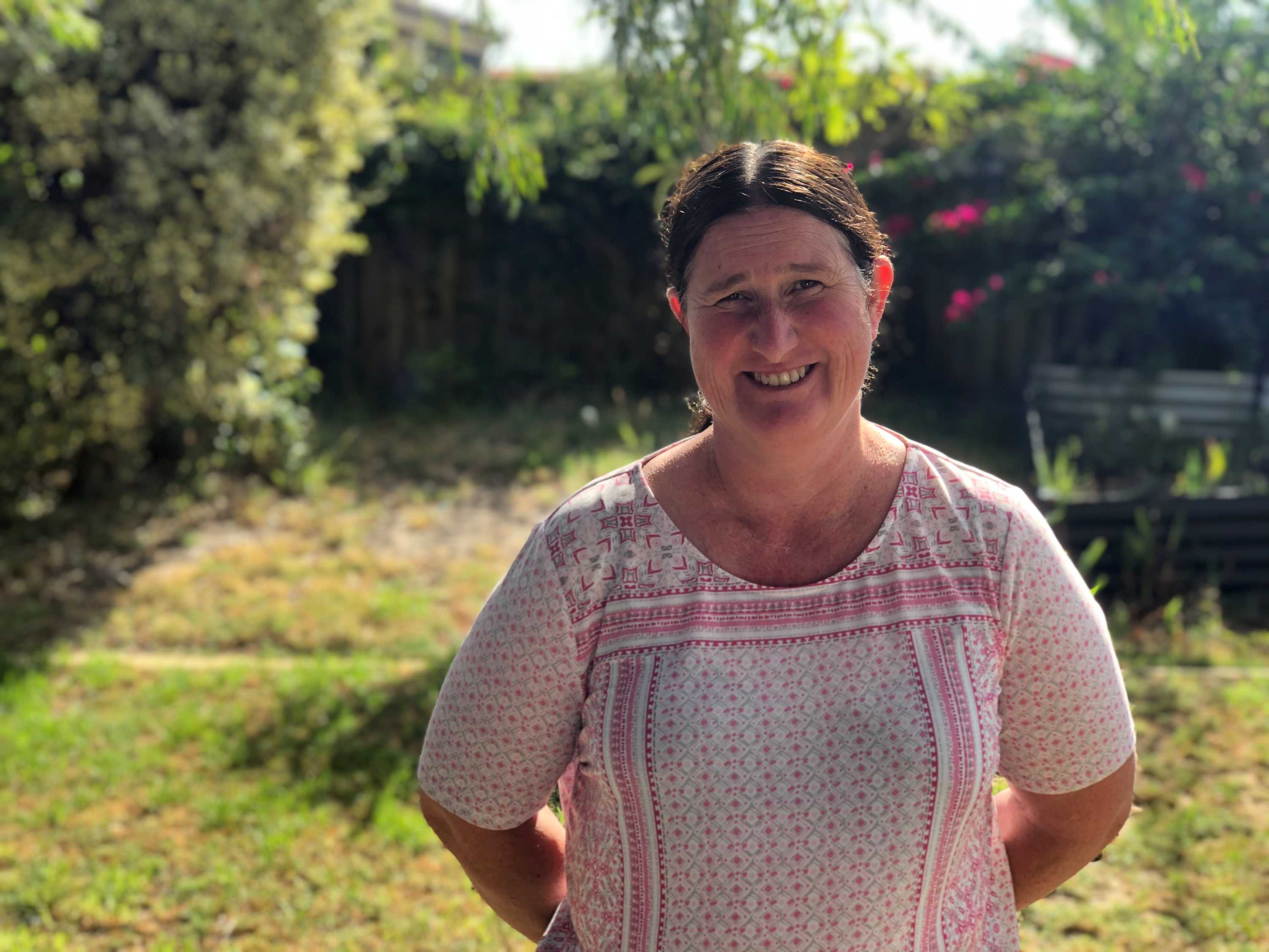 A woman wearing a pink and white shirt stands in a lush green backyard with grass and trees.