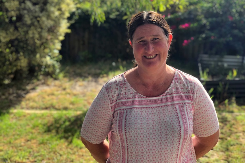 A woman wearing a pink and white shirt stands in a lush green backyard with grass and trees.