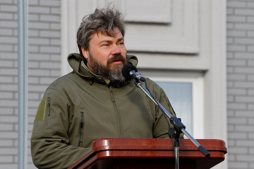 A heavyset middle-aged man with a dark beard wearing a brown jacket speaks outside behind a lectern.