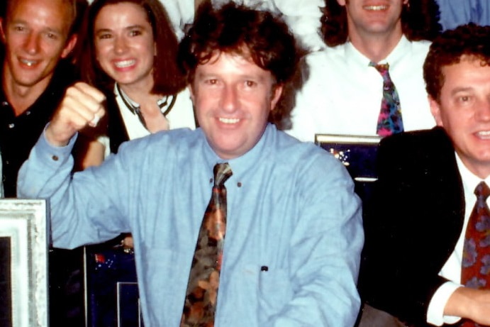 Denis Handlin smiles and holds up a fist while crouching in a group photo.