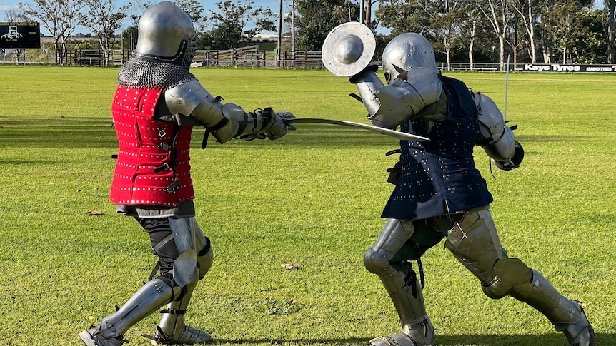 A man in armour &a red coat hits another man with a sword in full armour & a blue coat who is lunging with a small shield