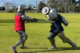 A man in armour &a red coat hits another man with a sword in full armour & a blue coat who is lunging with a small shield