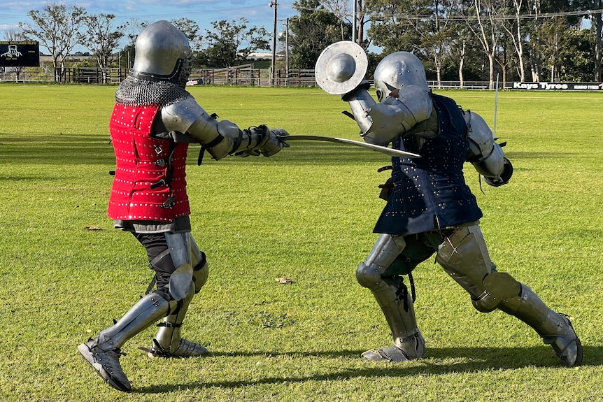 A man in armour &a red coat hits another man with a sword in full armour & a blue coat who is lunging with a small shield