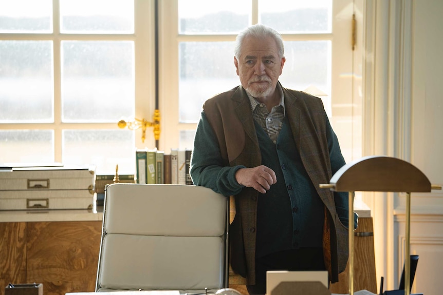 The actor stands next to a desk chair, his arm resting on the chair's back, in a brightly lit and well-decorated office.