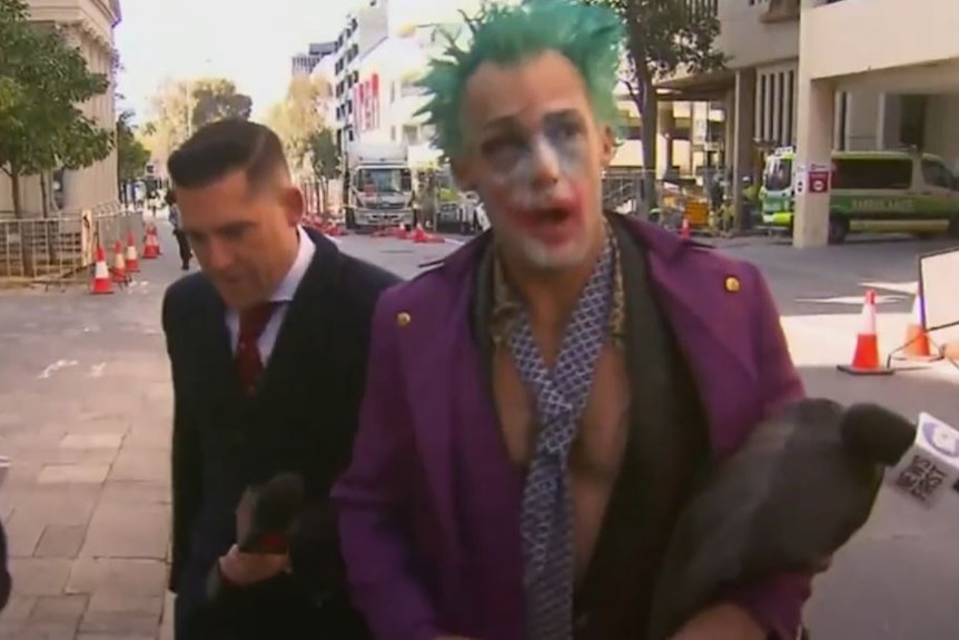 Reece Sturgeon, in full clown makeup, followed by a man in a suit, walks and speaks into a microphone.