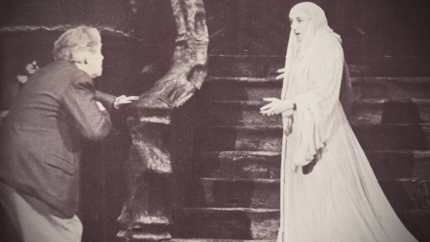 Sepia photo of man directing opera singer dressed in a white dress and veil on a stage. She is singing.