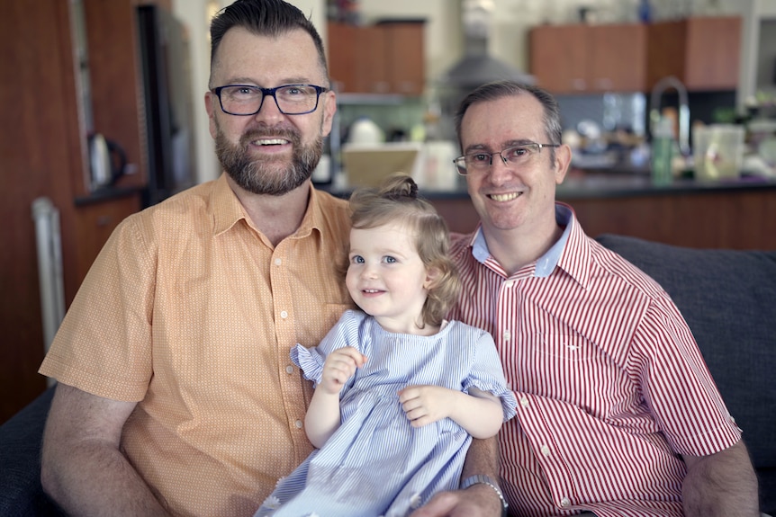 Two men smile holding their toddler daughter.