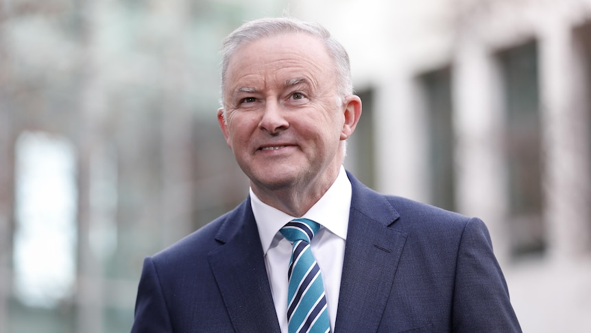 Albanese is outdoors, wearing a blue suit with blue and black tie, and smiling.