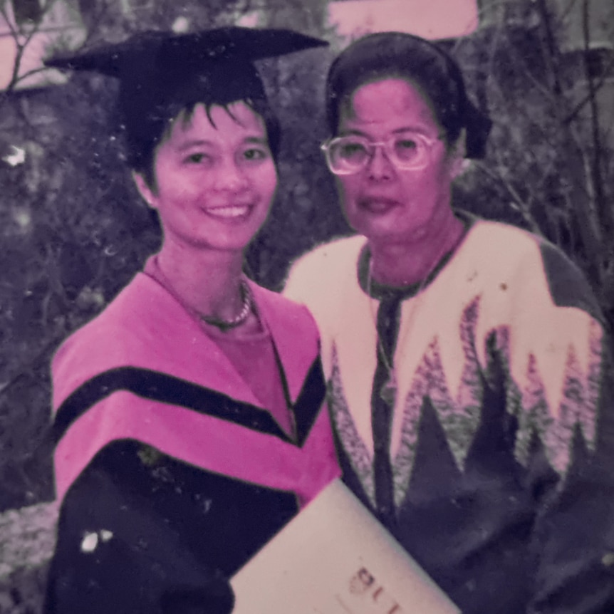 A young woman in a graduation gown stands next to an older woman