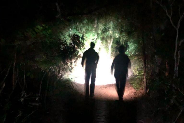 Two silhouettes with a torch on a path at night through the bush