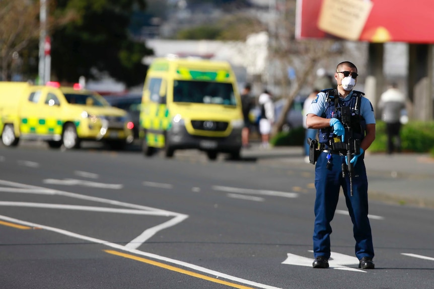 A police officer holding a machine gun stands on a road with ambulances behind him