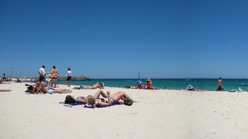 A wide panoramic shot of people sunbathing and swimming on Cottesloe beach in Perth under a bright blue sky.