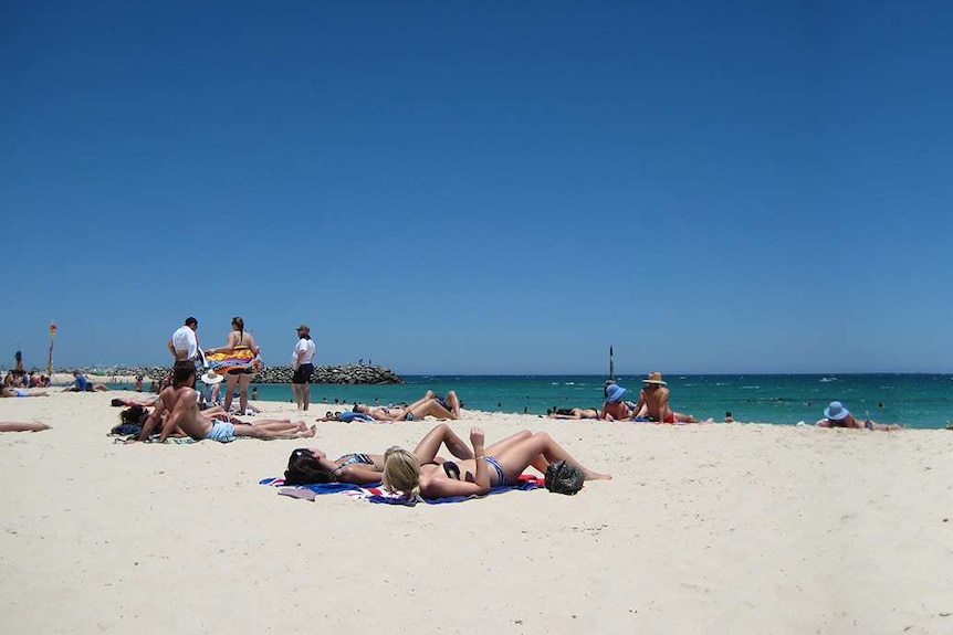 A wide panoramic shot of people sunbathing and swimming on Cottesloe beach in Perth under a bright blue sky.