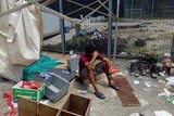 A man sits with his hand on his head, surrounding by trash and debris.