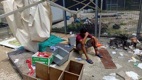 A man sits with his hand on his head, surrounding by trash and debris.